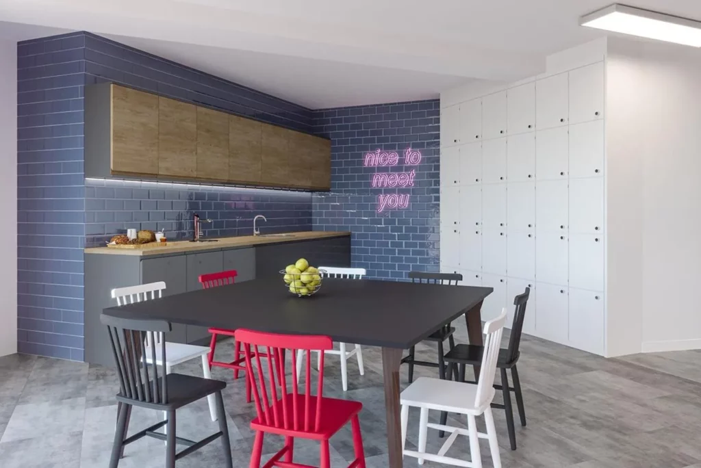 An office kitchen with lockers, a table and chair, kitchen worktops and a neon wall sign.