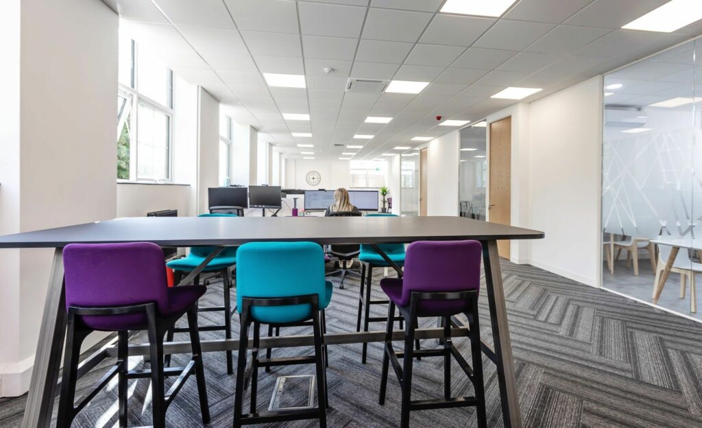 An office space with a tall table, high stools and desks in the background with separate offices on the right.