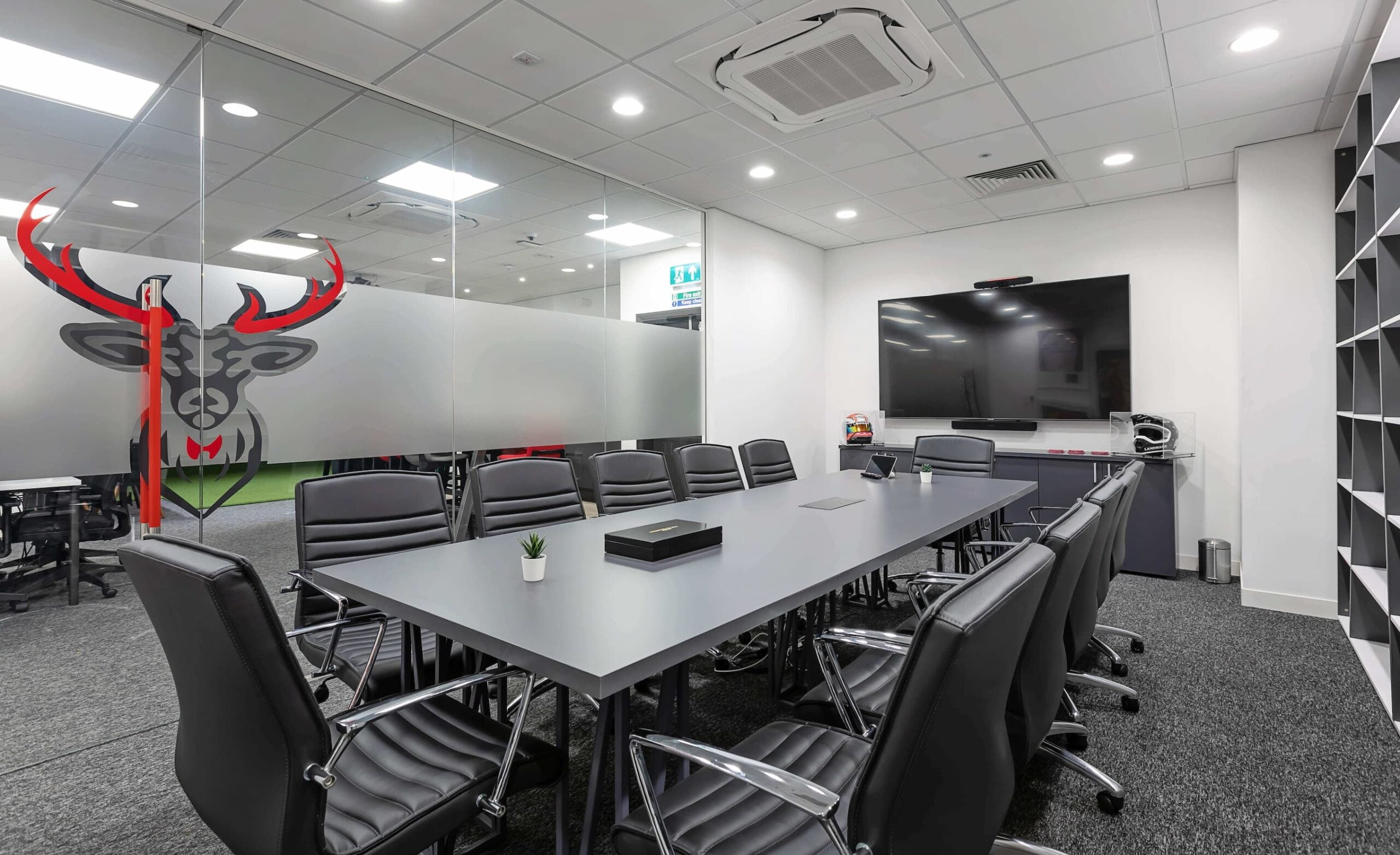 A bright meeting room with a dark grey table surrounded by chairs, a television mounted on the back wall and a glass tall with a stag design printed onto it.
