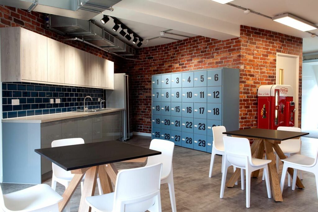 A kitchen area with blue tiles, blue lockers, two tables with chairs and a vintage coke machine.