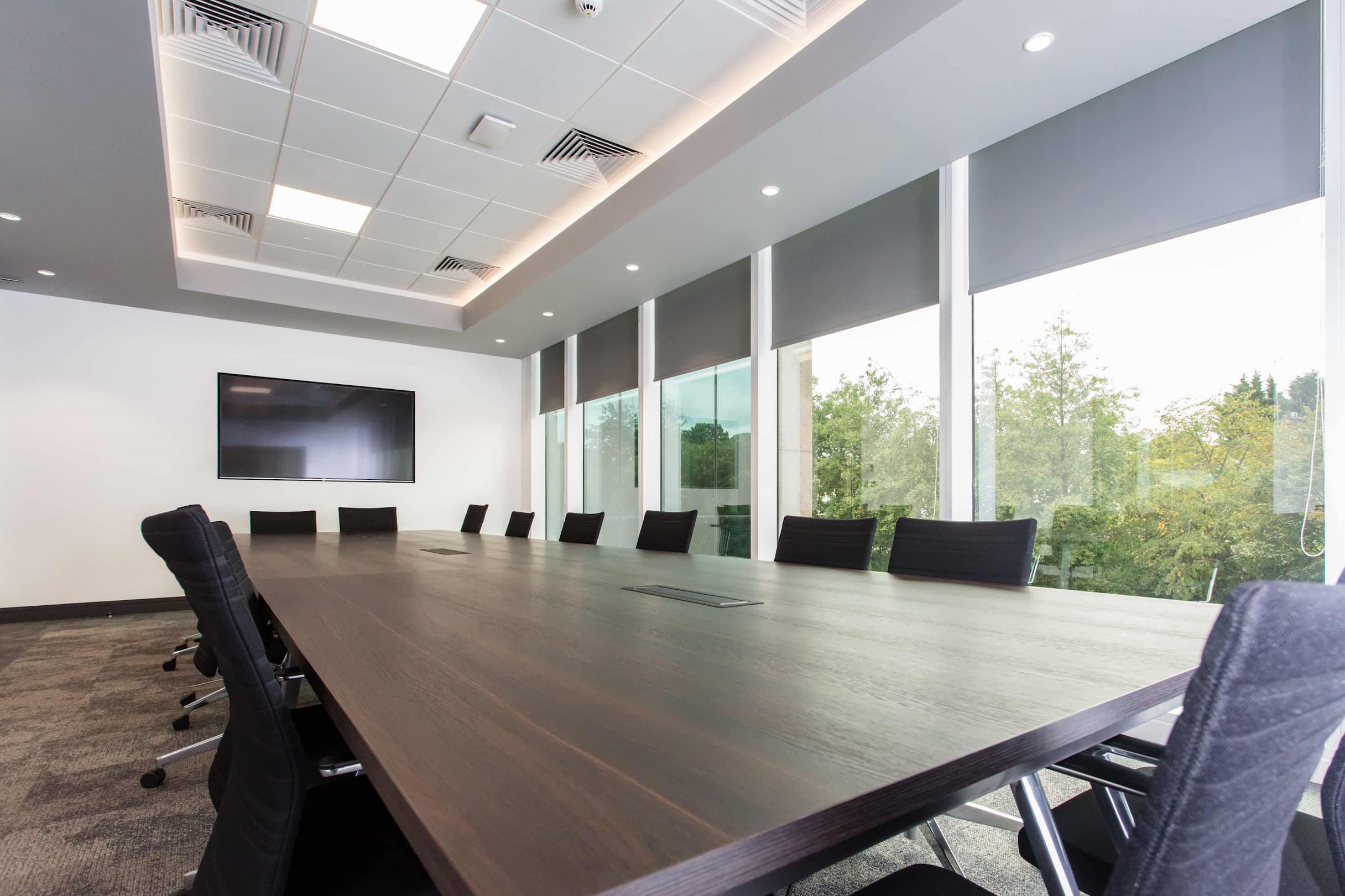 A meeting room with a dark wooden table, black chairs and windows on the right wall.