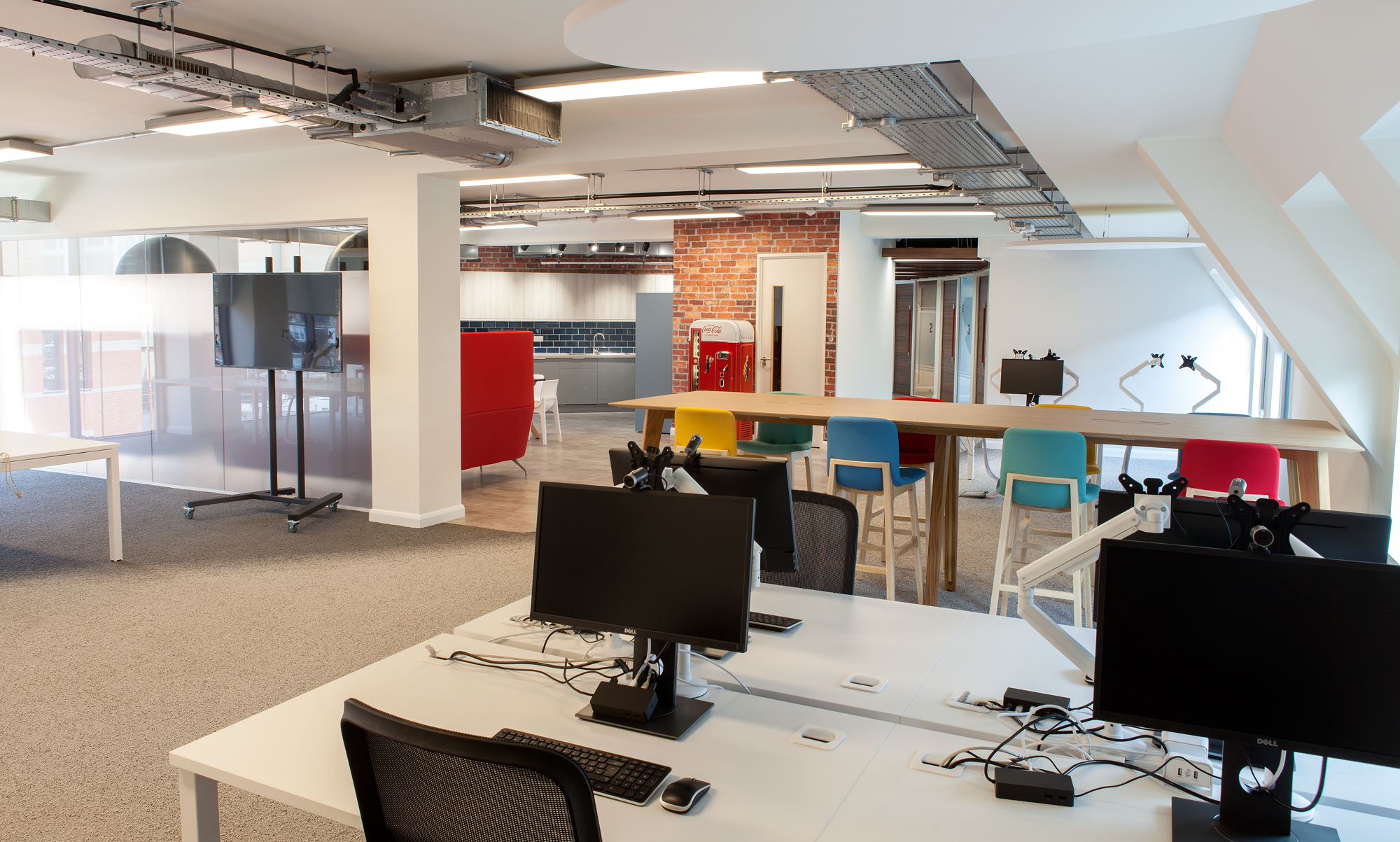 An office with desks and chairs, a television and a kitchen space in the background.